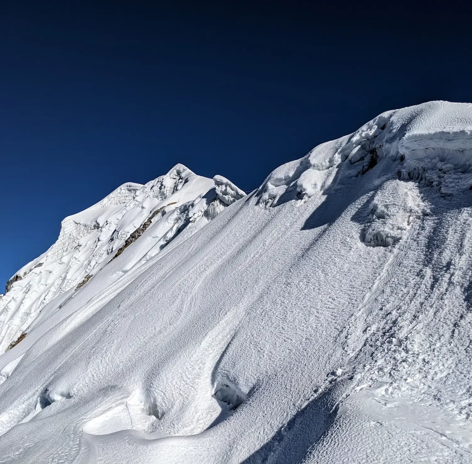 A steep glaciated summit ridge under a deep blue sky, high in the Nepal Himalaya